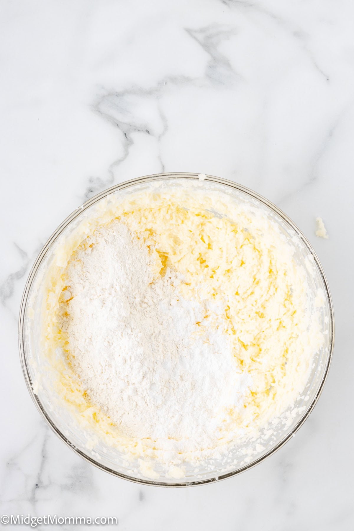 A glass bowl containing a mixture of white powdered sugar and yellow creamed butter on a white marble surface.