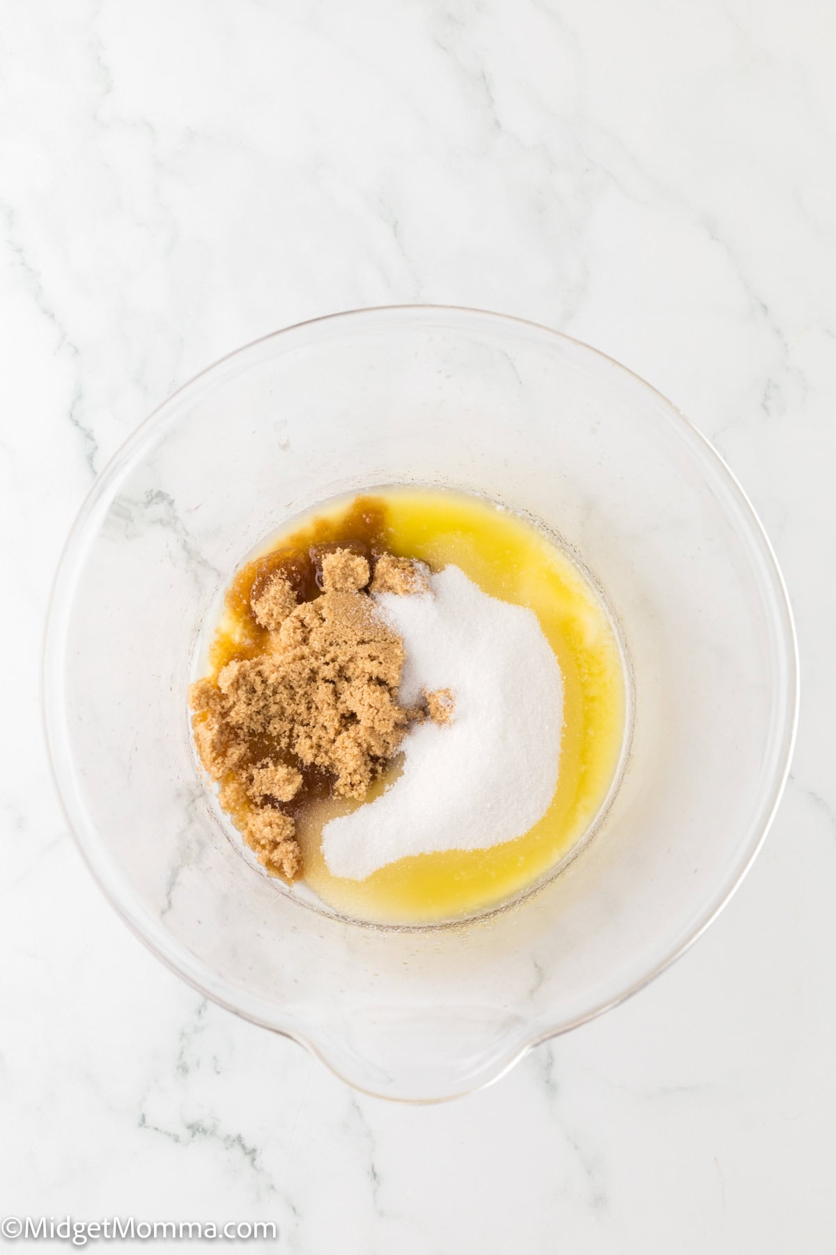 A clear glass bowl containing melted butter, brown sugar, and granulated white sugar on a white marble surface.