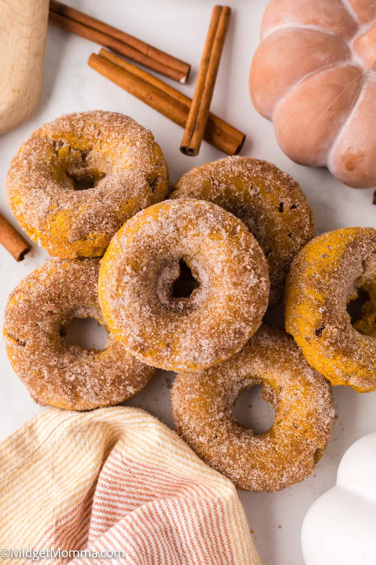 A group of sugar-coated pumpkin donuts arranged on a white surface with cinnamon sticks and decorative pumpkins nearby.