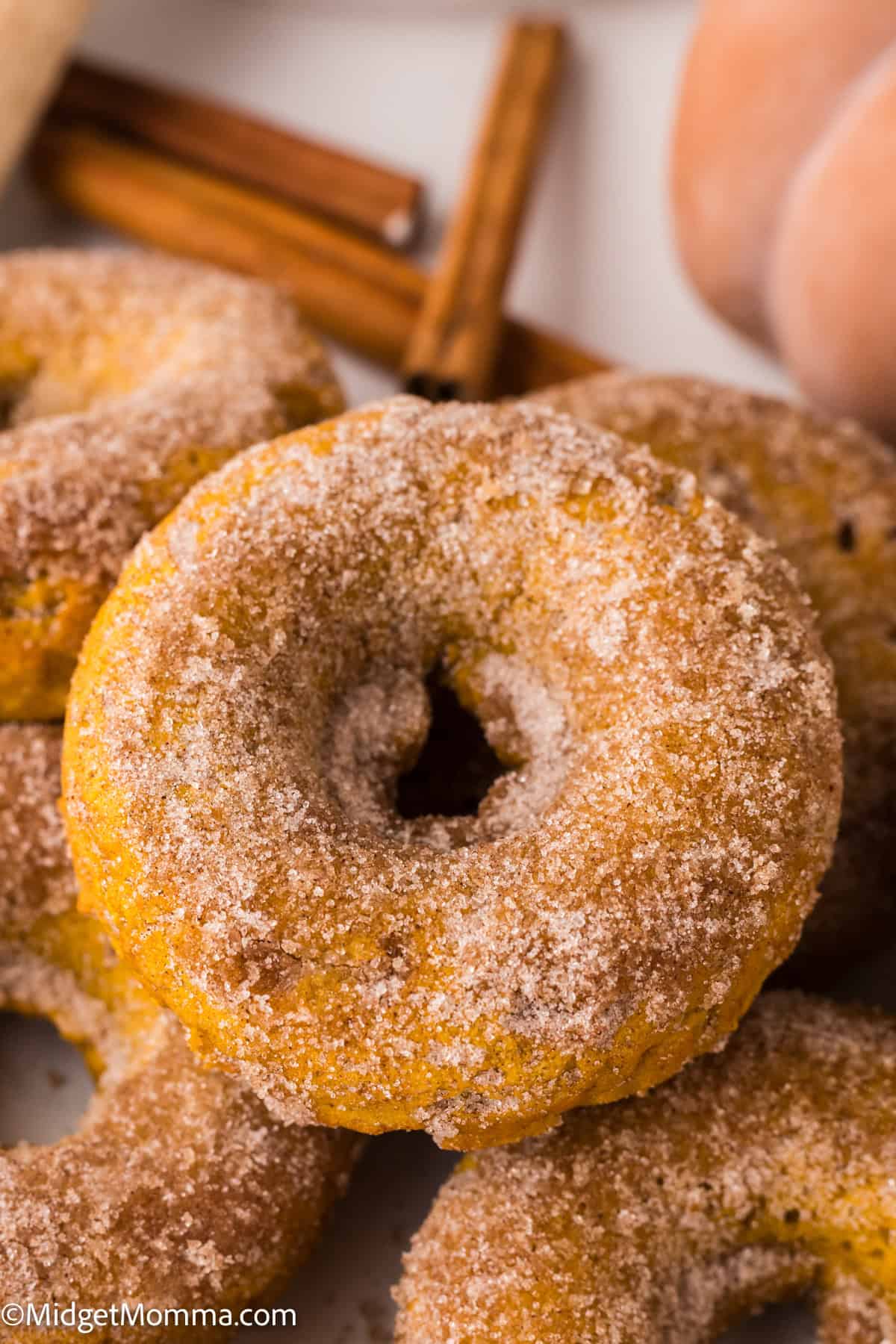 A close-up of sugar-coated baked pumpkin donuts stacked on a plate, with cinnamon sticks in the background.