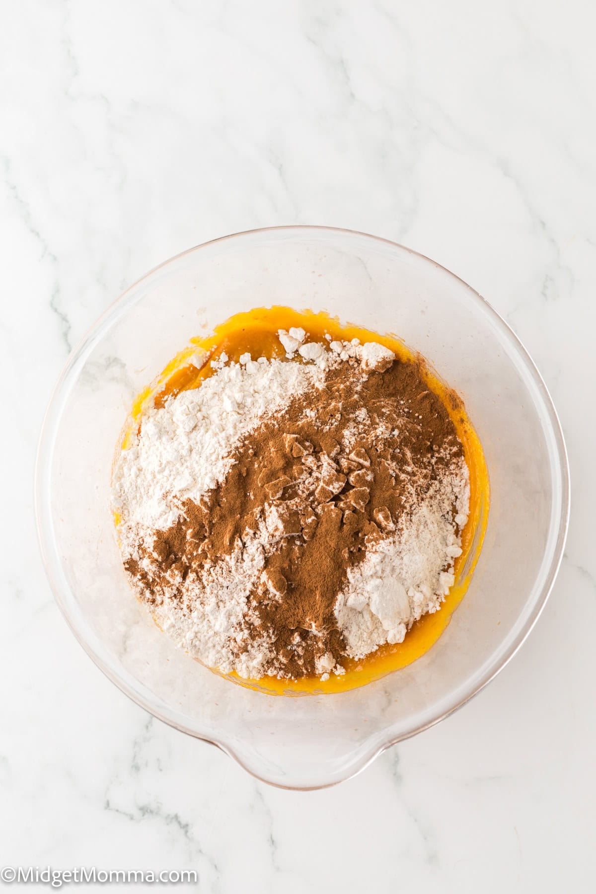 A glass mixing bowl containing flour, cinnamon, and a yellow batter mixture on a marble countertop.