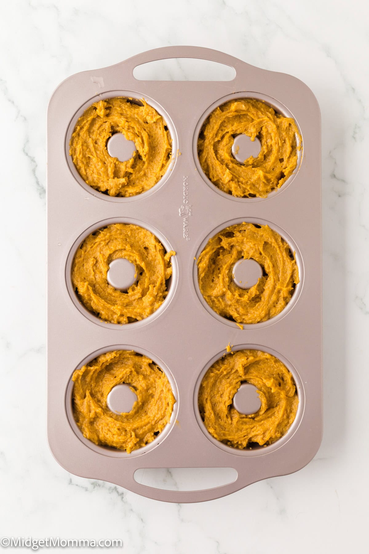 A 6-cavity donut pan filled with unbaked pumpkin batter sits on a white marble surface.