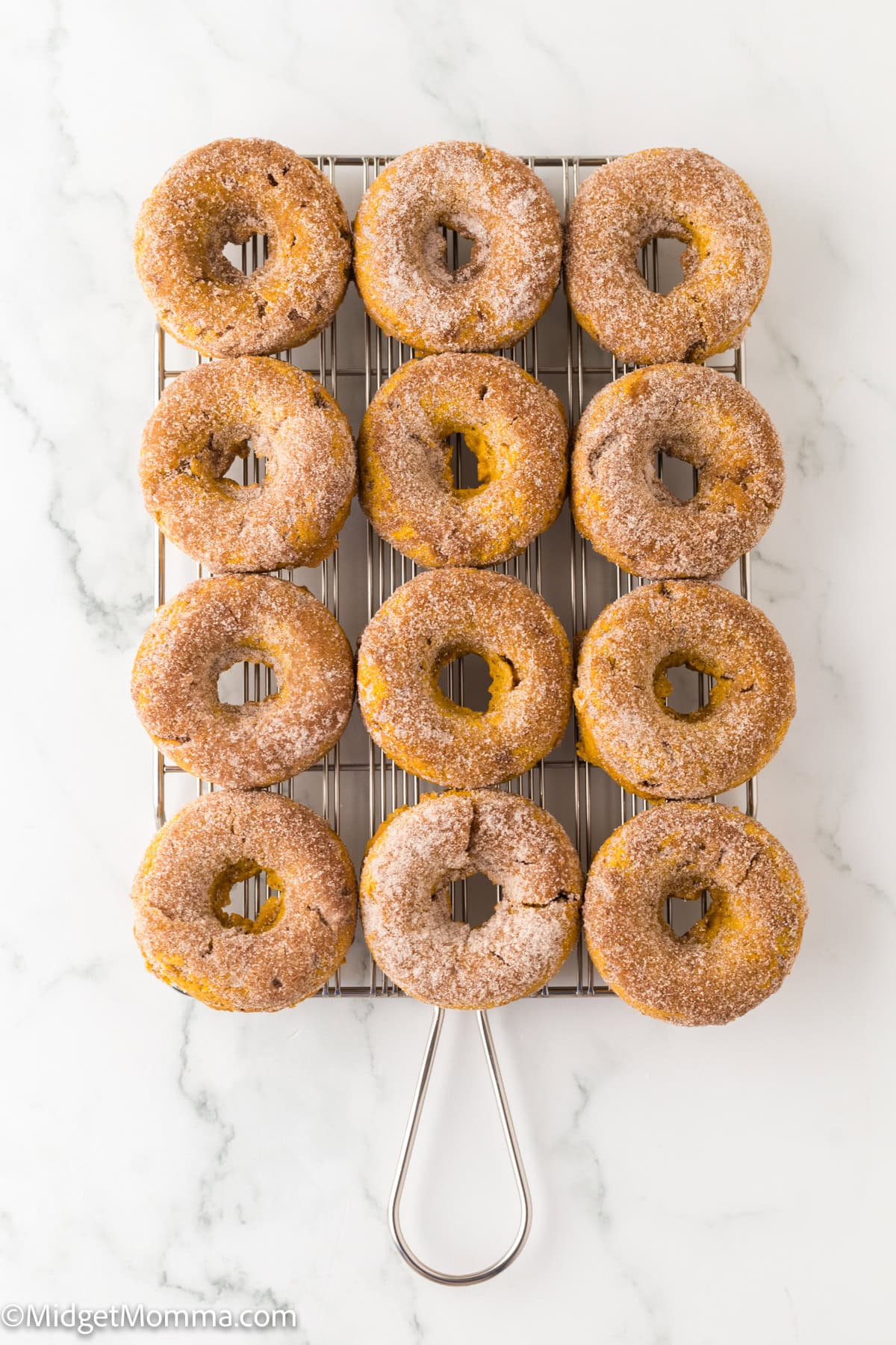 Twelve sugar-coated donuts are arranged in three rows on a cooling rack set on a white marble surface.