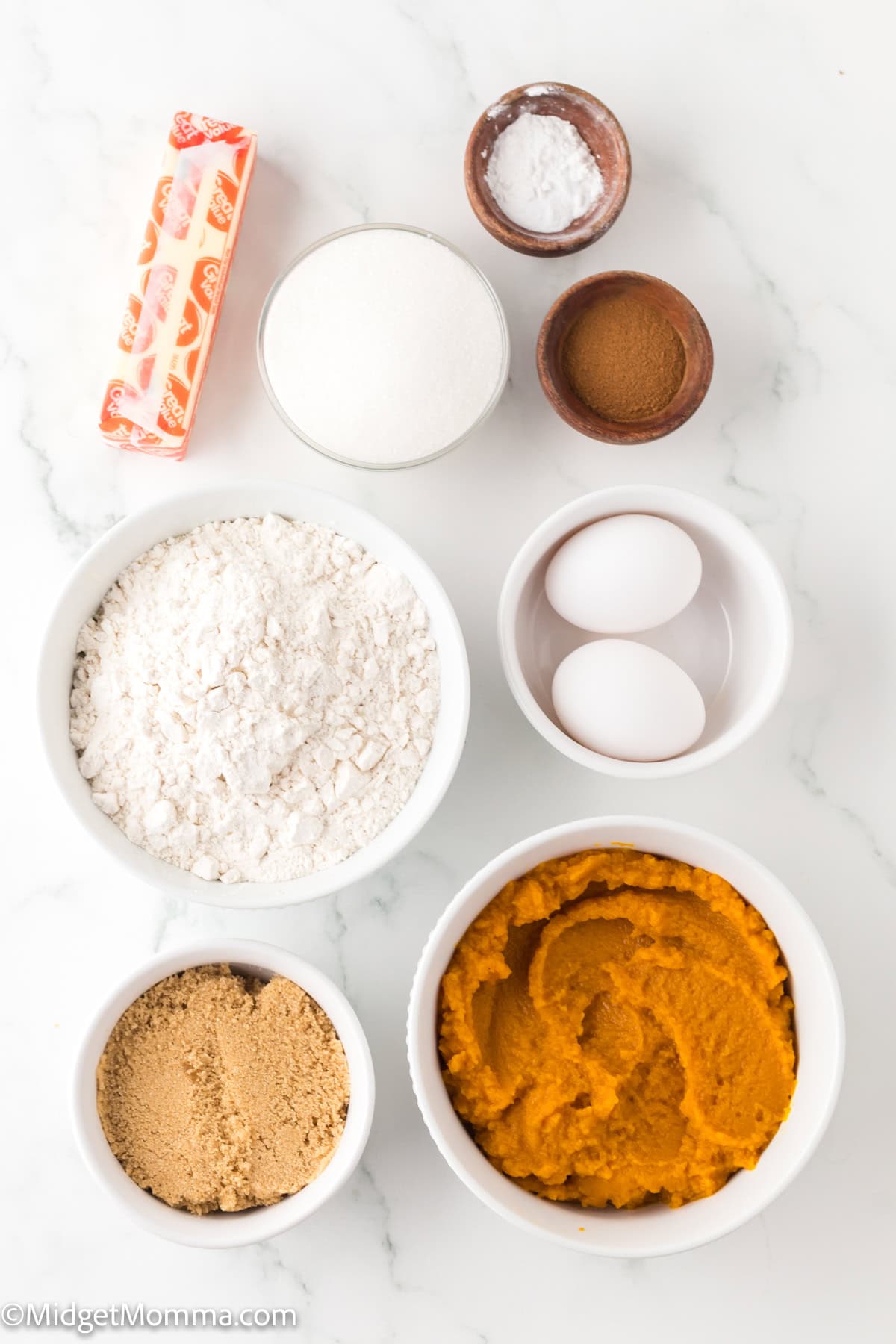 Baked Pumpkin Donuts Ingredients - Top-down view of baking ingredients, including butter, granulated sugar, brown sugar, flour, eggs, pumpkin puree, cinnamon, and baking powder, arranged in bowls on a white surface.