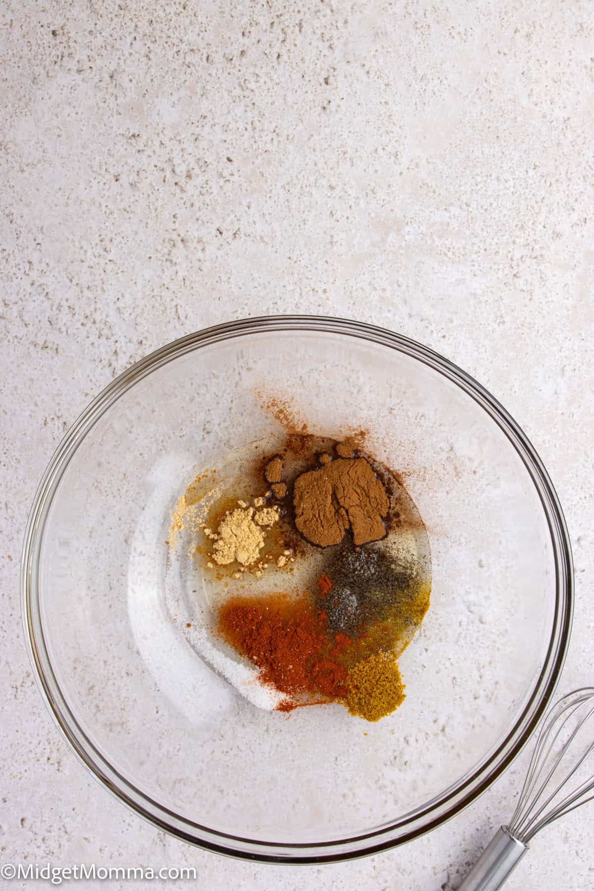 A glass mixing bowl containing various spices and sugar, placed on a light countertop with a metal whisk nearby.
