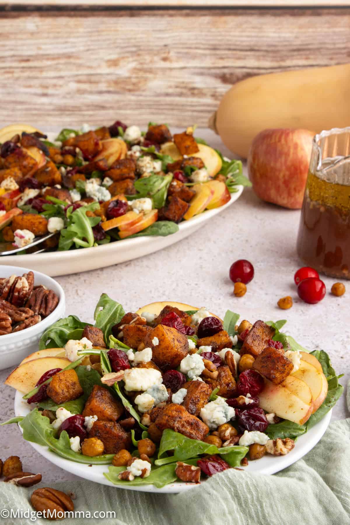A plate of autumn salad with apples, croutons, leafy greens, nuts, cranberries, blue cheese, and a jar of vinaigrette on the side.