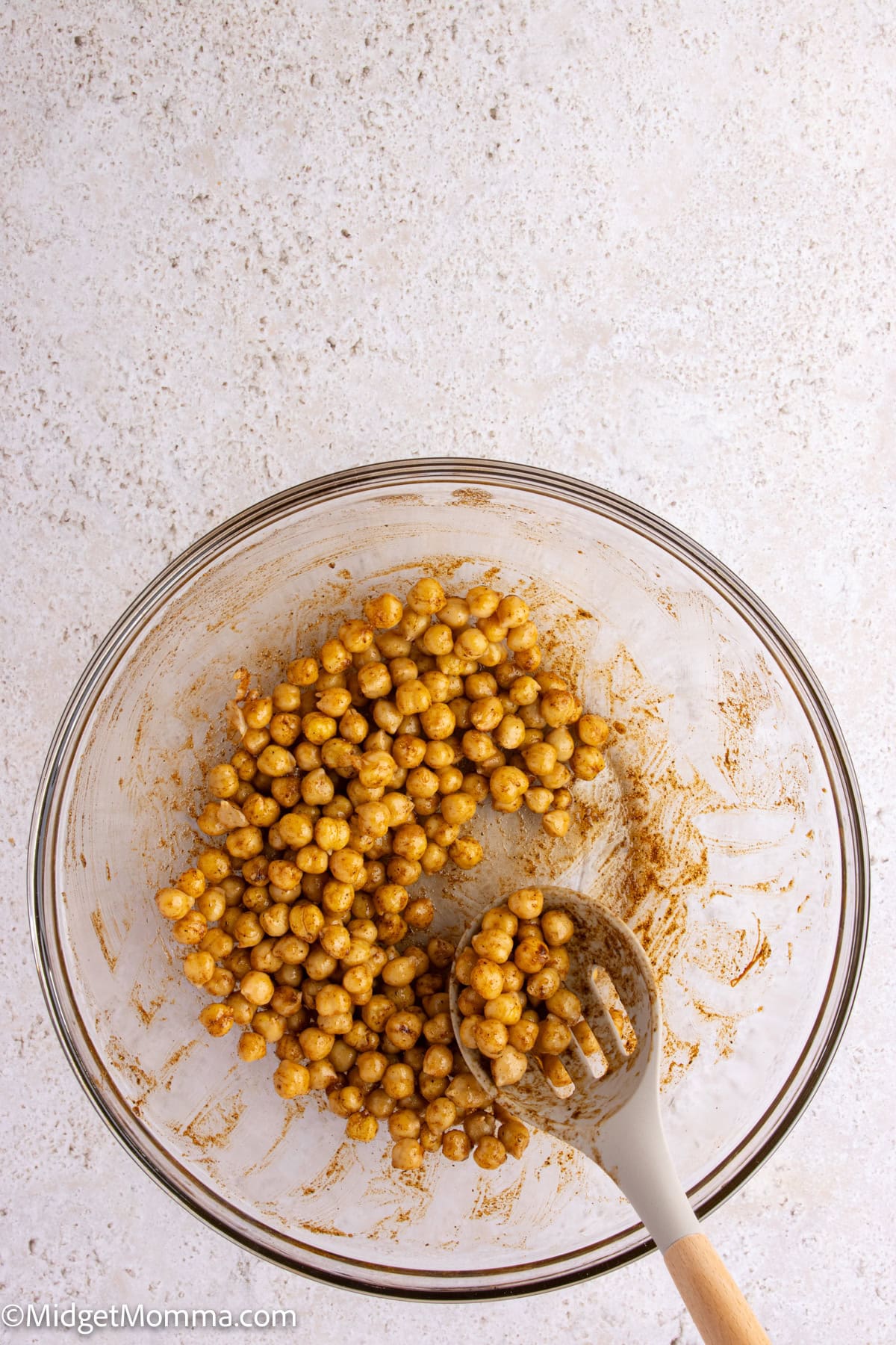 A glass bowl containing seasoned chickpeas with a slotted spoon resting inside on a light-colored surface.
