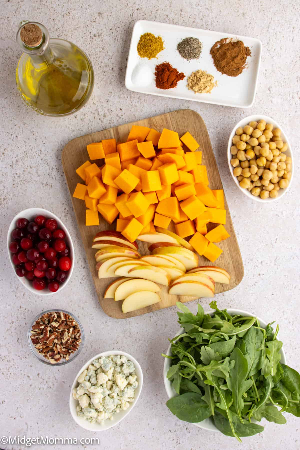 Overhead view of ingredients including cubed butternut squash, sliced apples, arugula, chickpeas, cranberries, pecans, crumbled cheese, spices, and a bottle of olive oil on a counter.