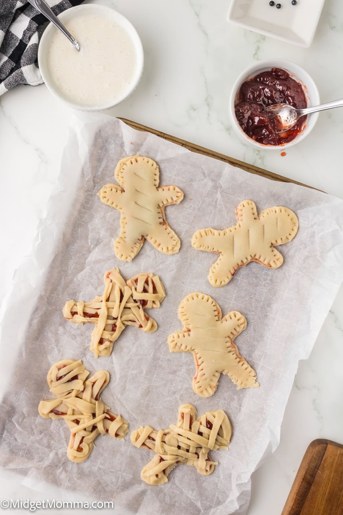 A baking tray with unbaked gingerbread-shaped pastries filled with jam, some with lattice tops, sits on parchment paper next to bowls of sugar and jam.