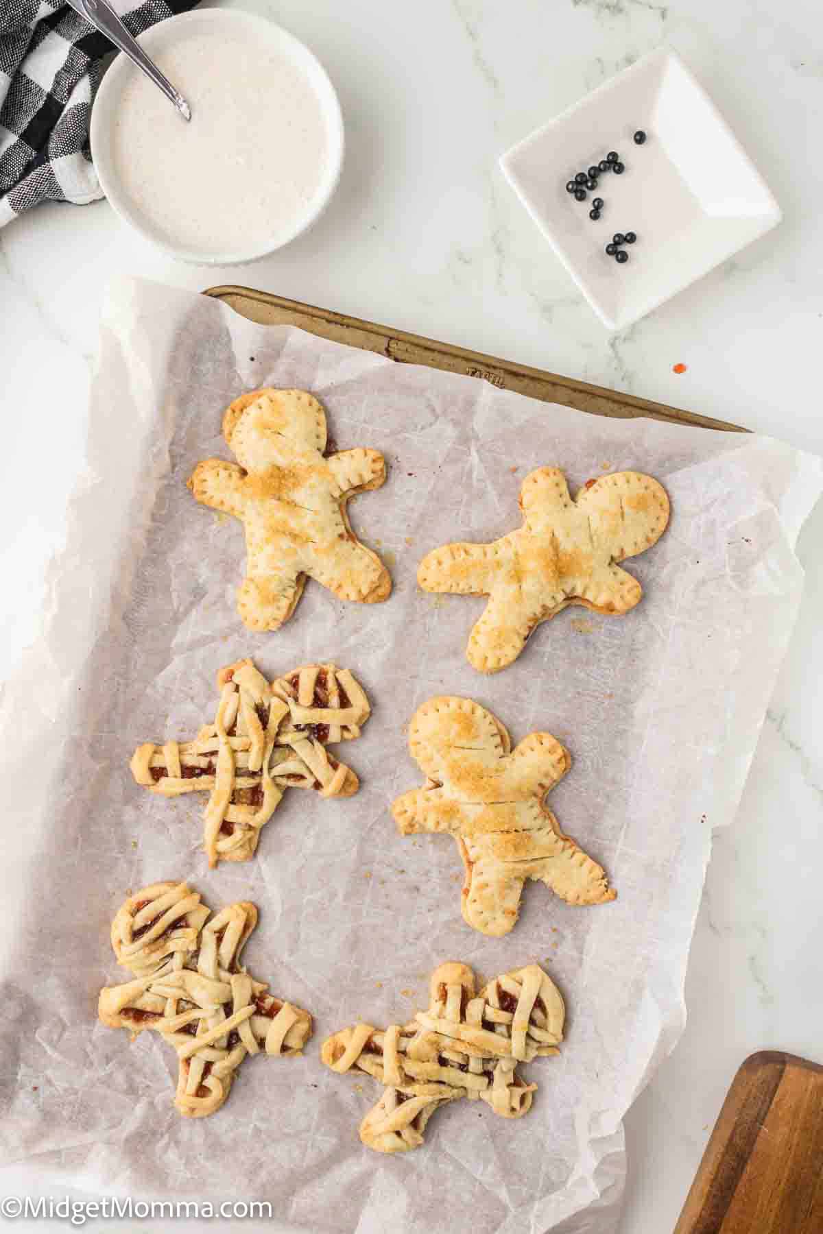 A baking sheet with six gingerbread man-shaped pastries, some with a lattice pattern, next to a bowl of white glaze and a dish of black decorations.
