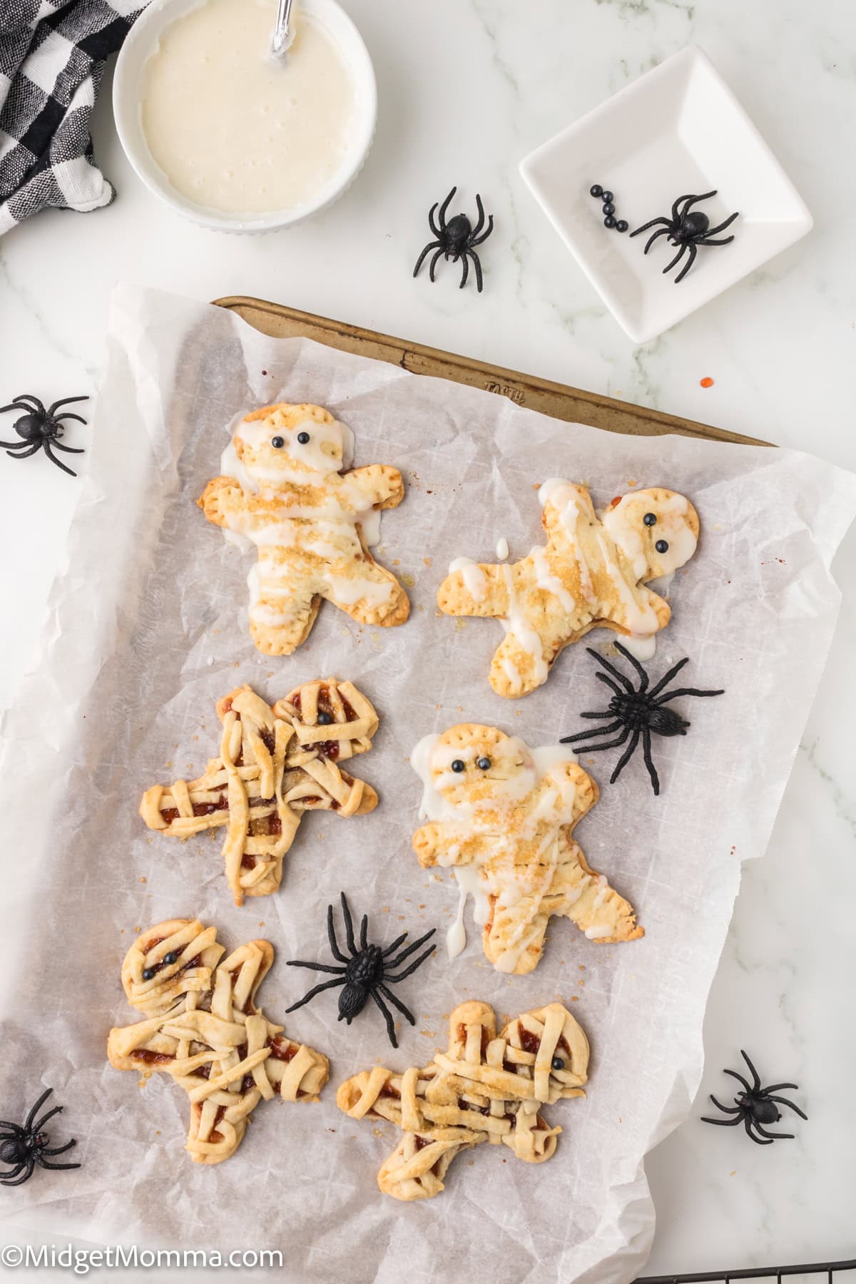 A baking sheet with six mummy-shaped pastries, some with white icing and some with a lattice pattern, surrounded by plastic black spider decorations on a white surface.