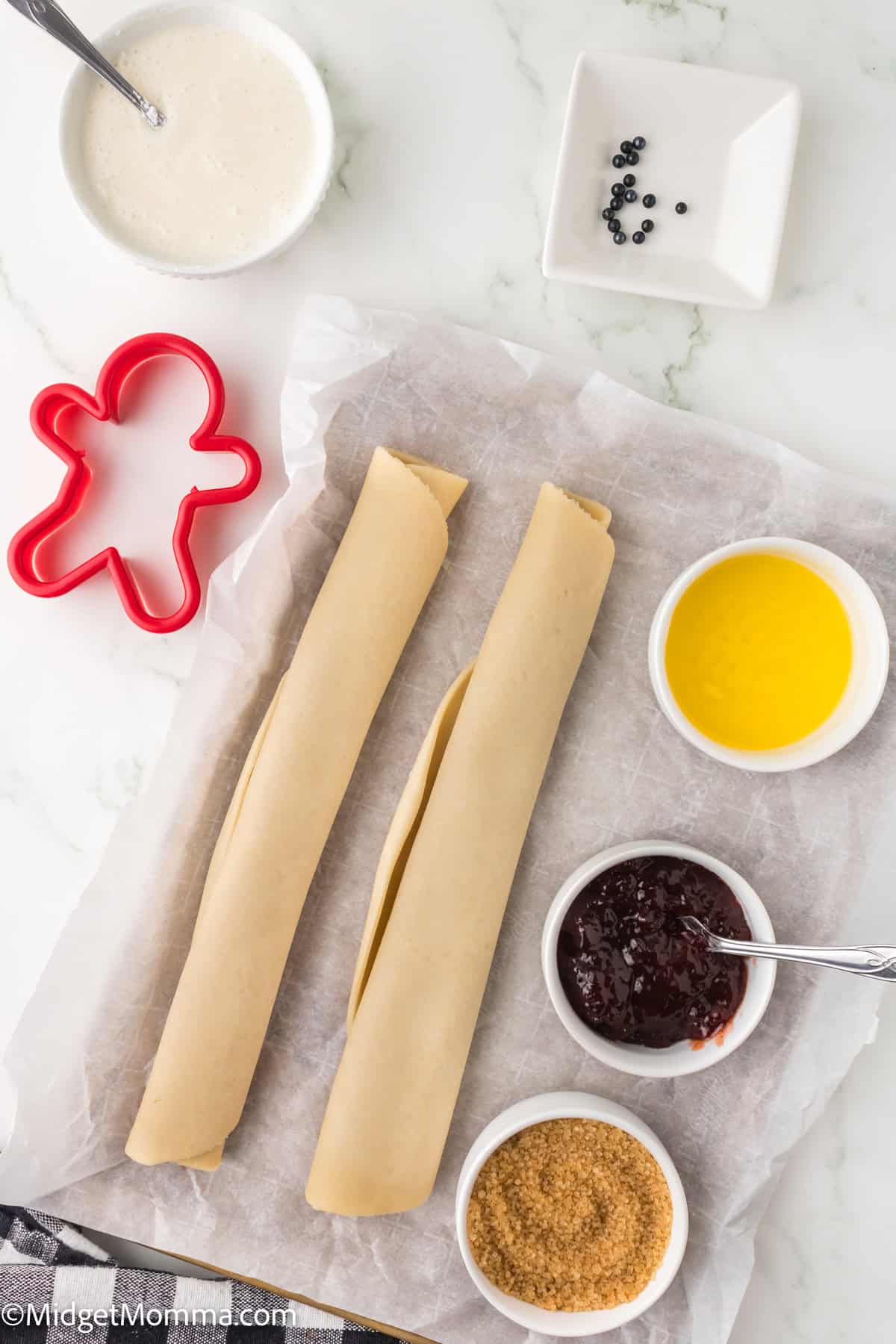 Two rolled dough sheets on parchment surrounded by bowls of powdered sugar glaze, melted butter, fruit jam, brown sugar, black sprinkles, and a red gingerbread man cookie cutter.