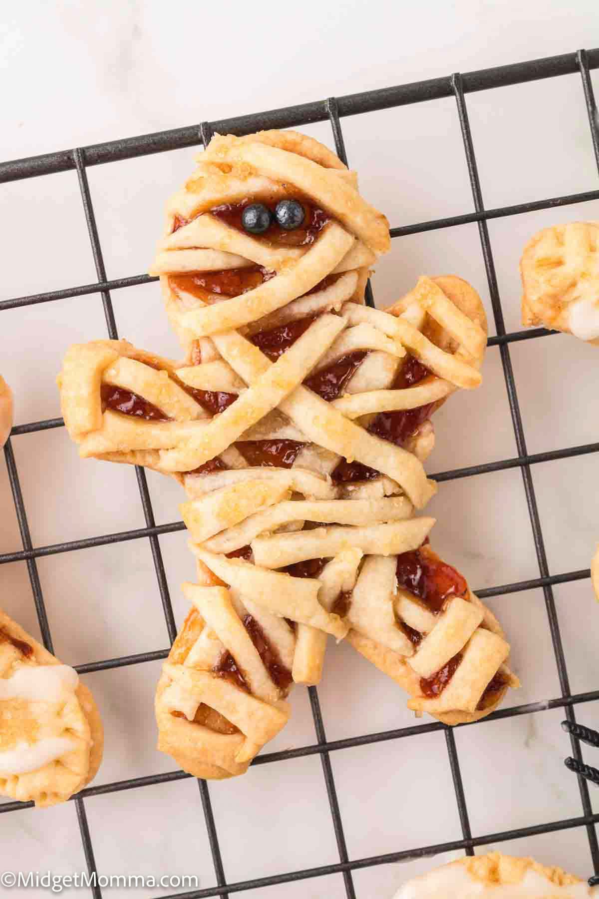 A cookie decorated to look like a mummy with pastry strips and two small black candy eyes, placed on a cooling rack.