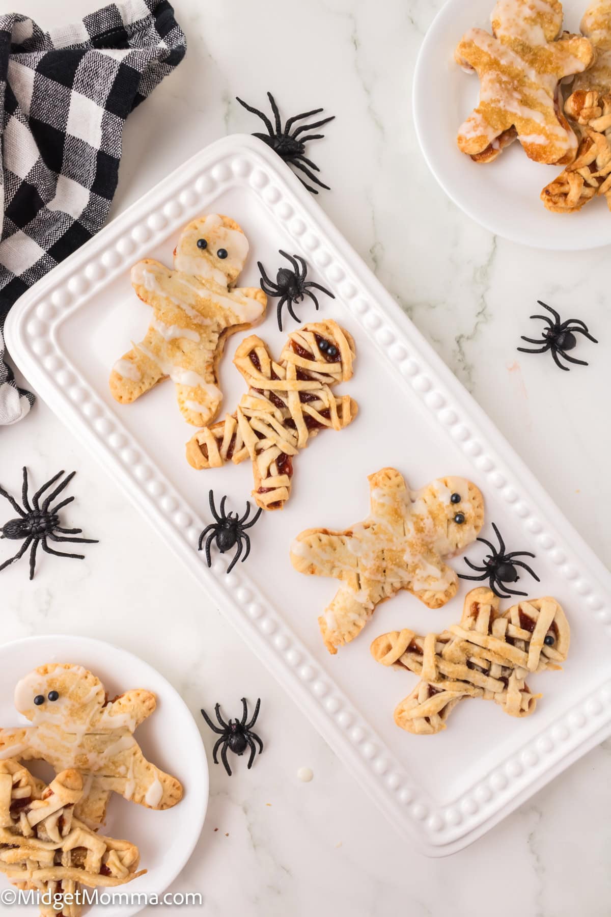 Rectangular white platter with Halloween-themed pastries shaped like mummies and gingerbread men, surrounded by fake black spiders on a marble surface.