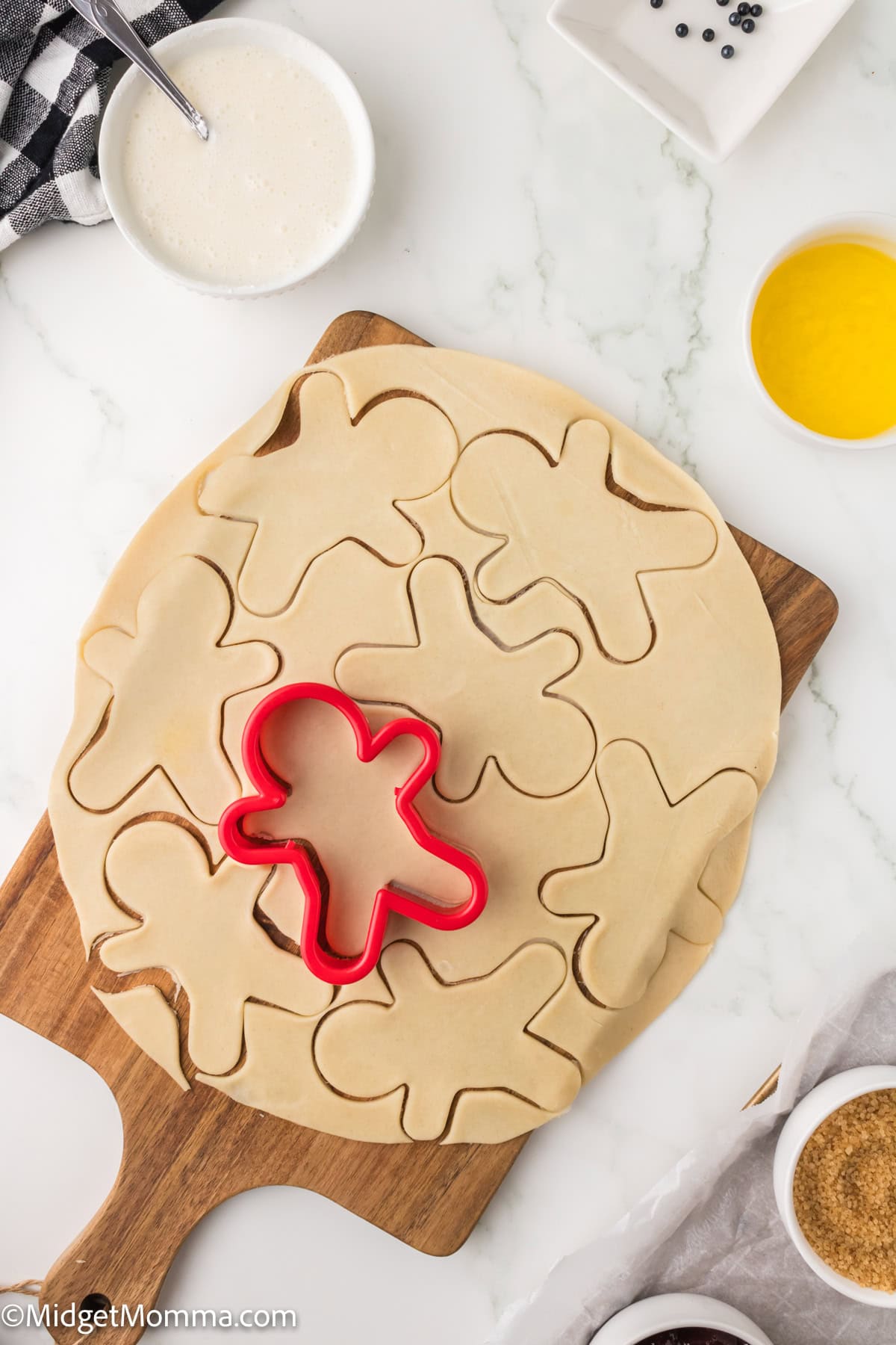A sheet of rolled dough with gingerbread person shapes cut out, a red cookie cutter, and baking ingredients on a white surface.