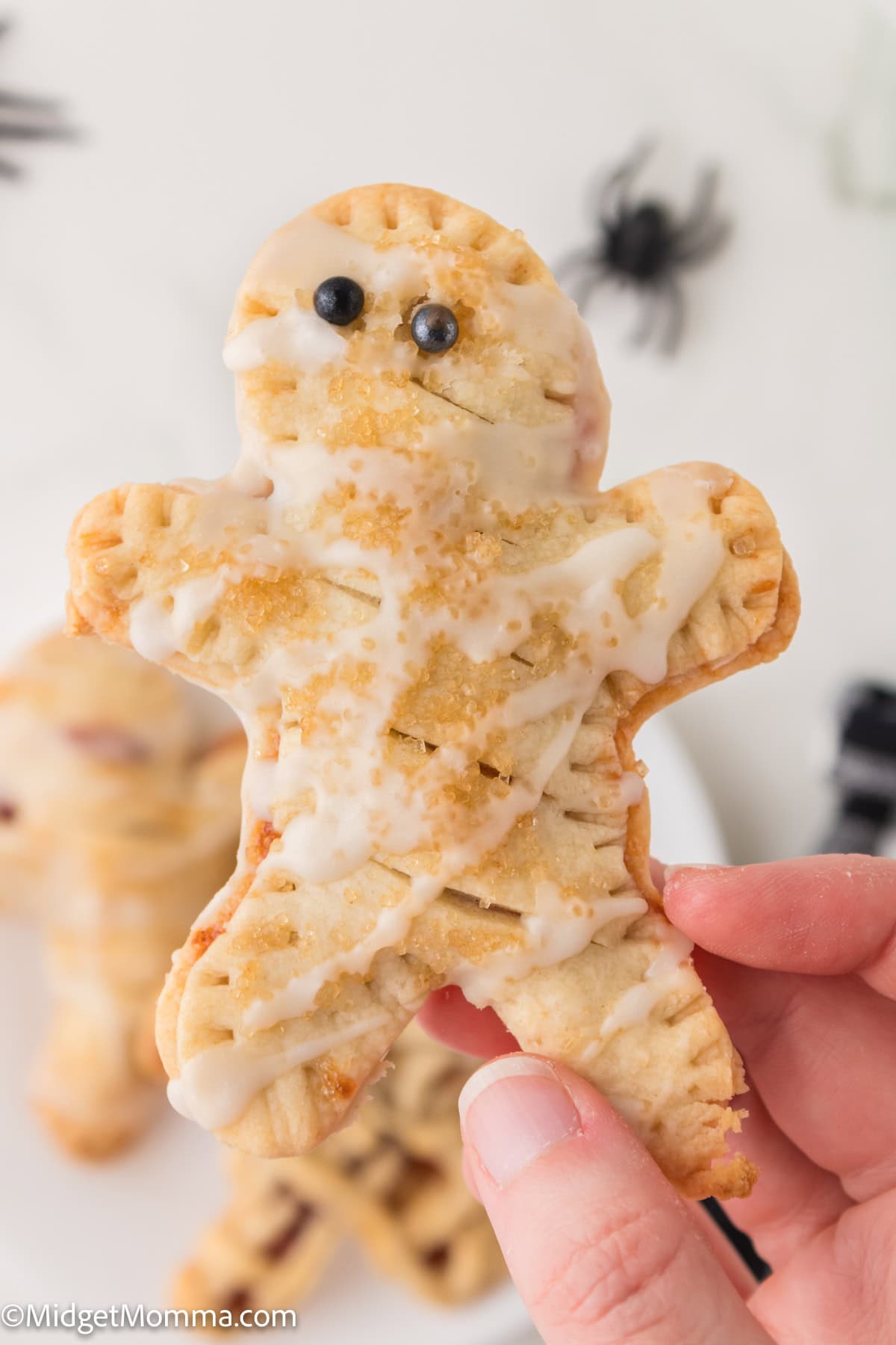 A hand holds a mummy-shaped pastry with icing and candy eyes; more pastries and plastic spiders are visible in the background.