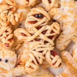 Several mummy-shaped cookies with icing and jam filling, decorated with strips to resemble mummy wrappings and small round candies for eyes, are arranged on a plate.