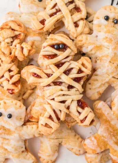 Several mummy-shaped cookies with icing and jam filling, decorated with strips to resemble mummy wrappings and small round candies for eyes, are arranged on a plate.