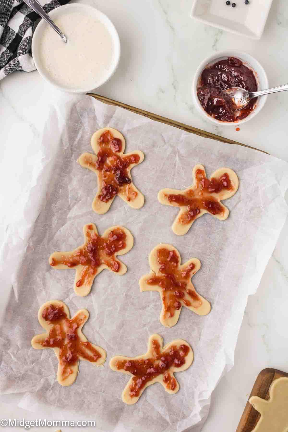 Six gingerbread-shaped cookies on a baking sheet are topped with red jam, with bowls of icing and jam nearby on a white countertop.