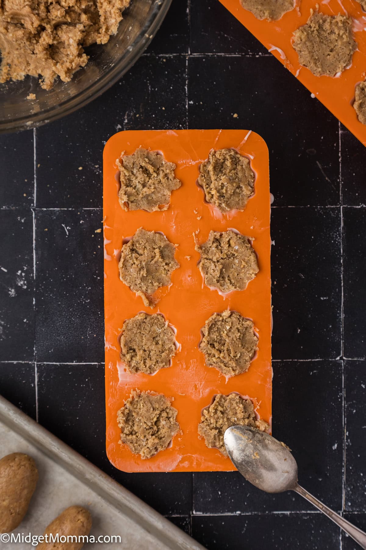 An orange silicone mold filled with scoops of brown dough sits on a black surface, with a spoon and a mixing bowl nearby.
