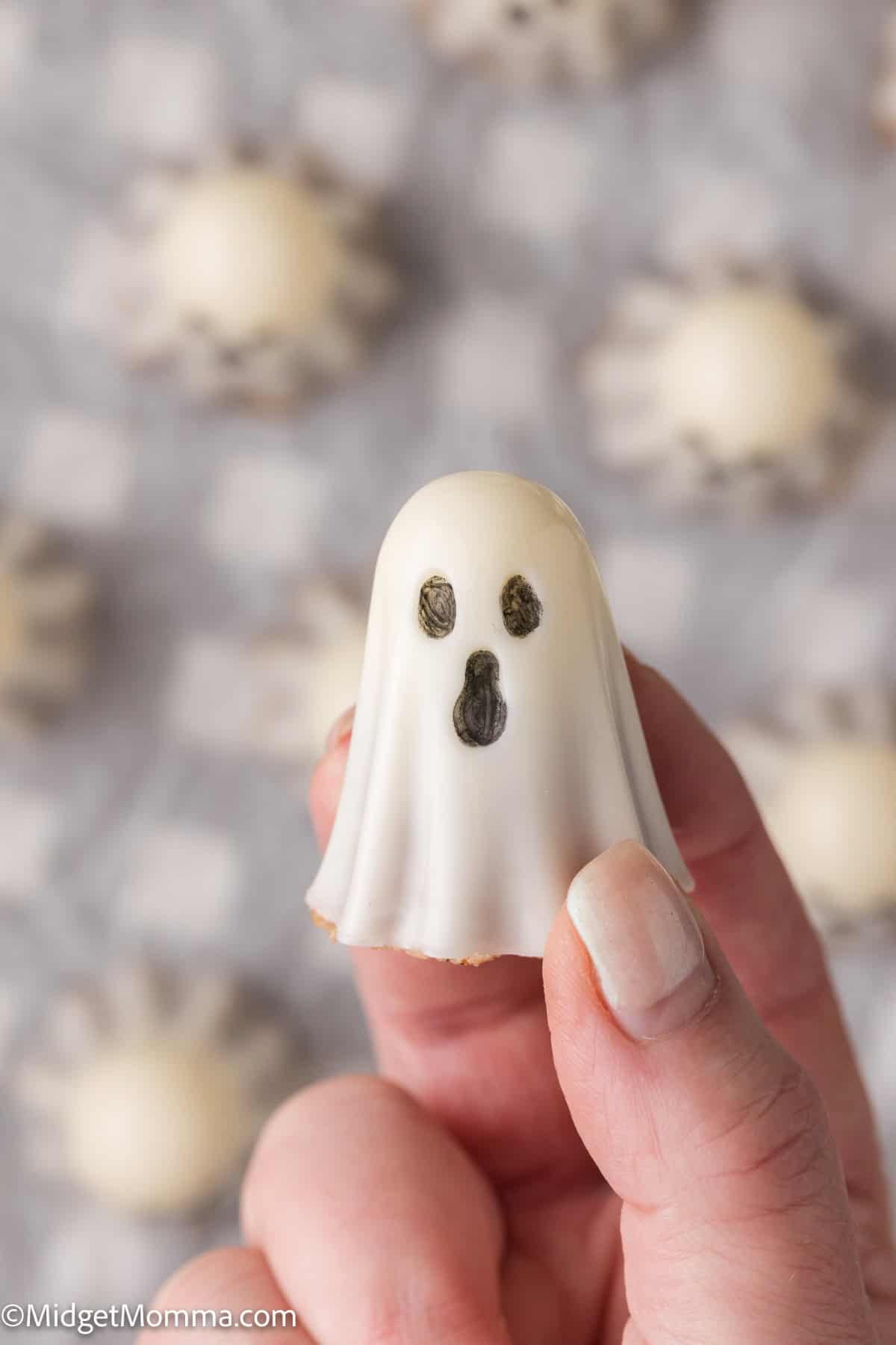A hand holding a small, white, ghost-shaped treat with dark eyes and mouth; blurred similar treats are in the background.