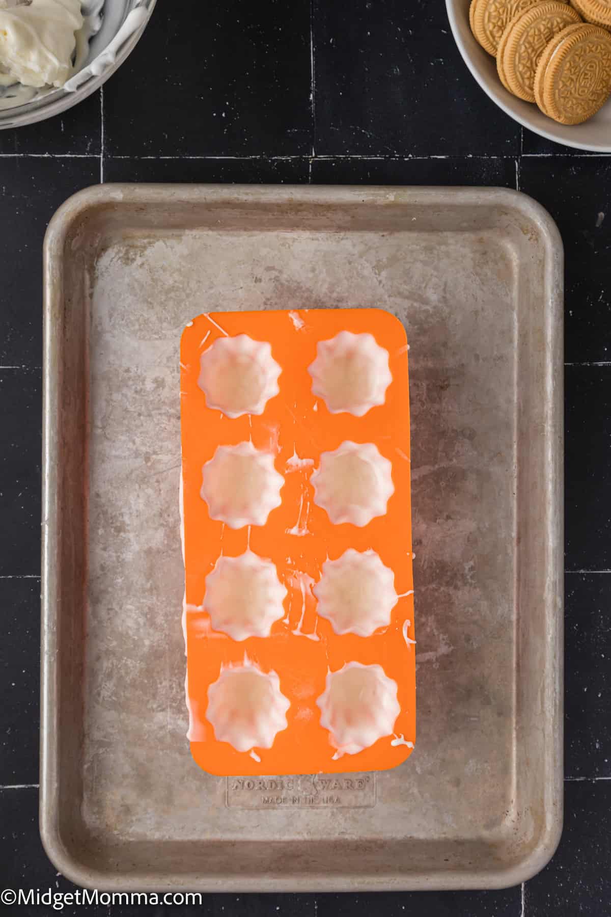 An orange silicone mold with eight white chocolate fillings sits on a metal baking sheet. A bowl of cream and a plate of cookies are nearby.