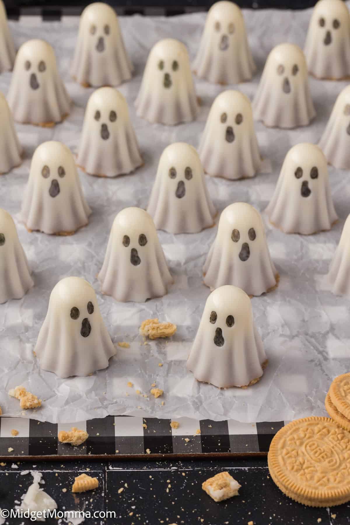 Rows of ghost-shaped white chocolate treats with cookie bases and chocolate eyes and mouths are arranged on wax paper, with cookie crumbs scattered nearby.