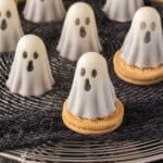 Several ghost-shaped white chocolate treats with dark eyes and mouths sit on round cookies, displayed on a wire rack with a black cloth underneath.
