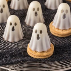 Several ghost-shaped white chocolate treats with dark eyes and mouths sit on round cookies, displayed on a wire rack with a black cloth underneath.