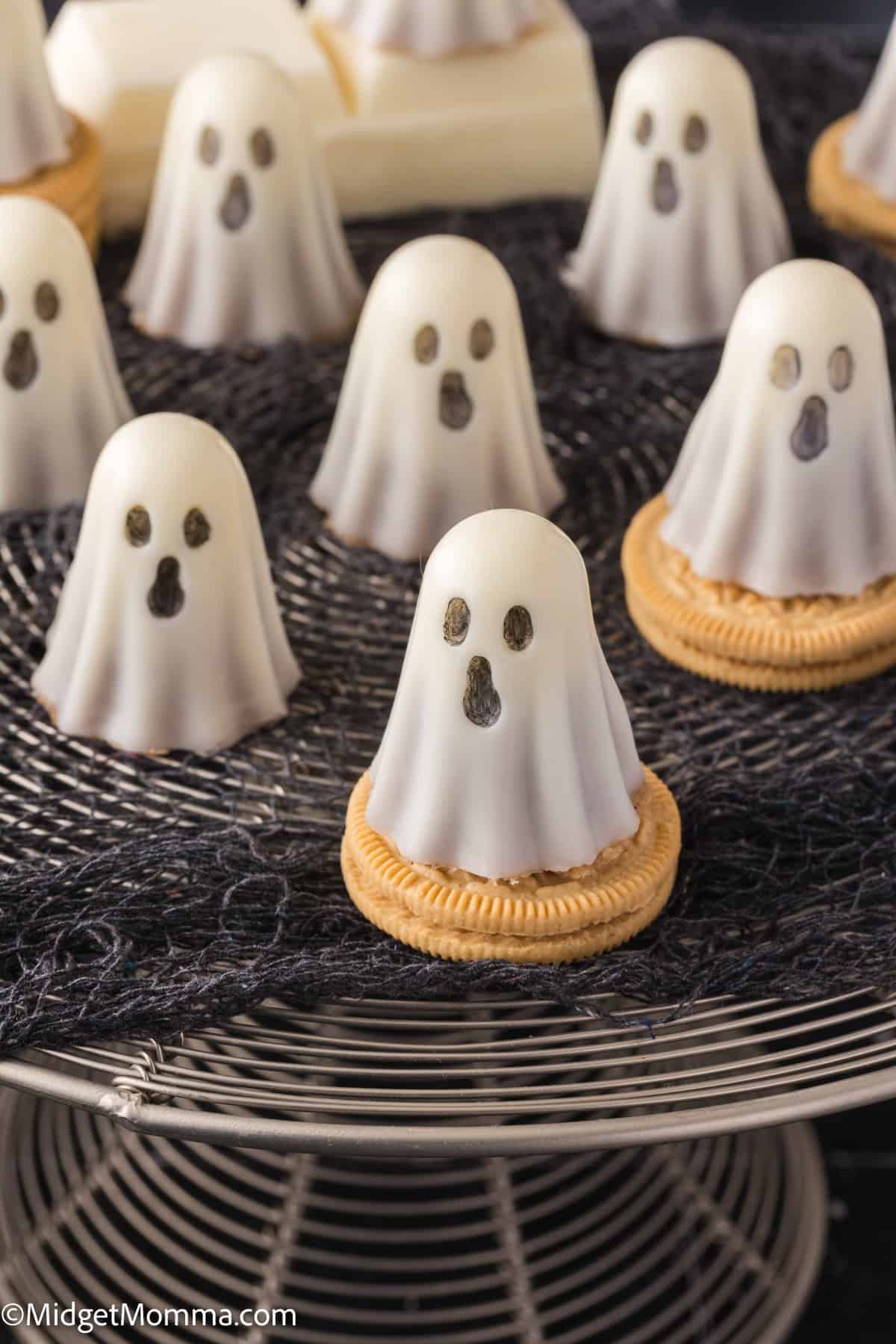 Several ghost-shaped white chocolate treats with dark eyes and mouths sit on round cookies, displayed on a wire rack with a black cloth underneath.