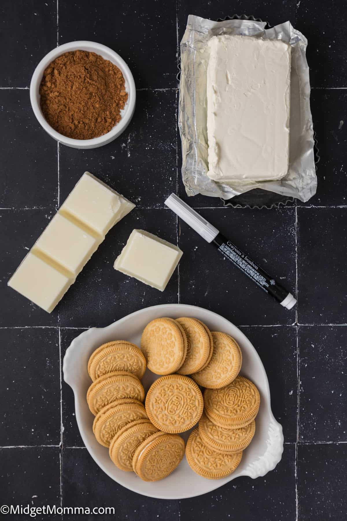 A bowl of golden sandwich cookies, a plate of white chocolate blocks, a dish of cocoa powder, a block of cream cheese, and a black edible marker on a black tiled surface.