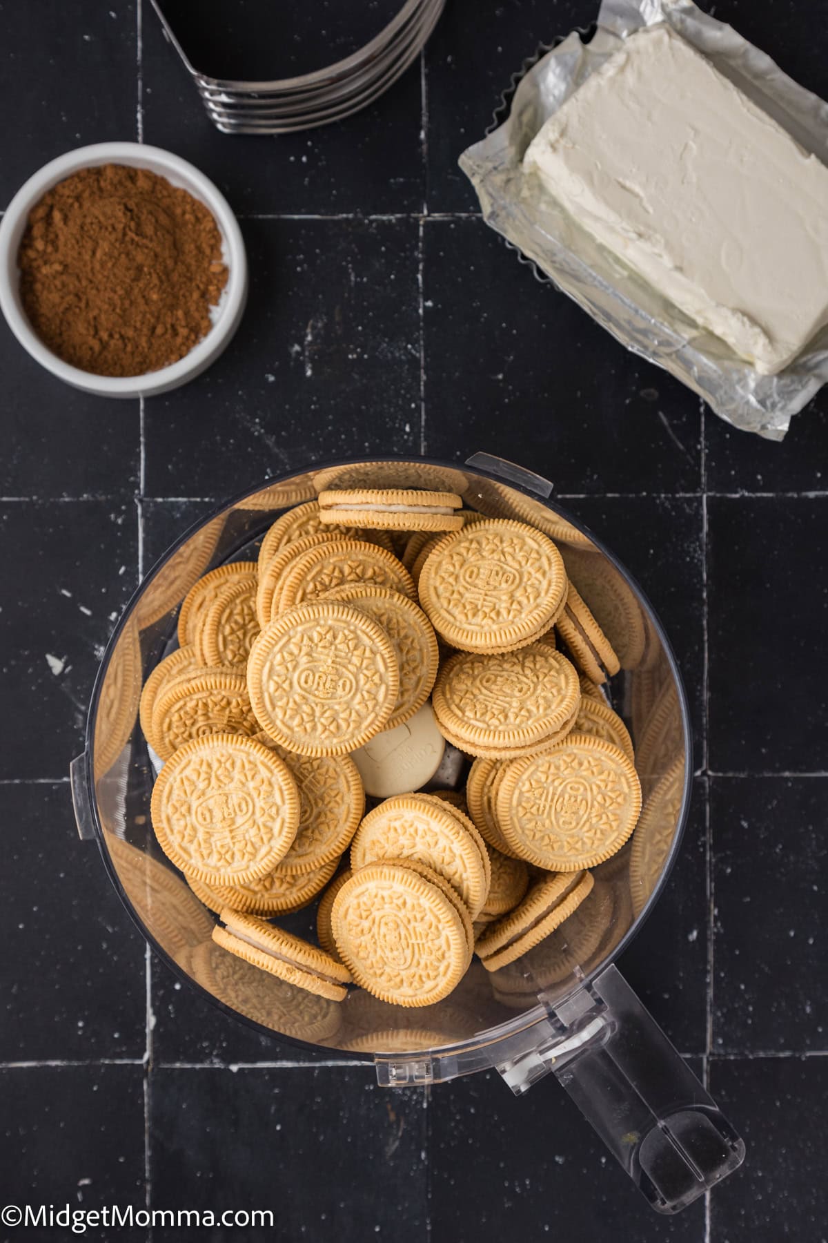 A food processor bowl filled with golden sandwich cookies sits on a black tiled surface next to a block of cream cheese and a small bowl of cocoa powder.