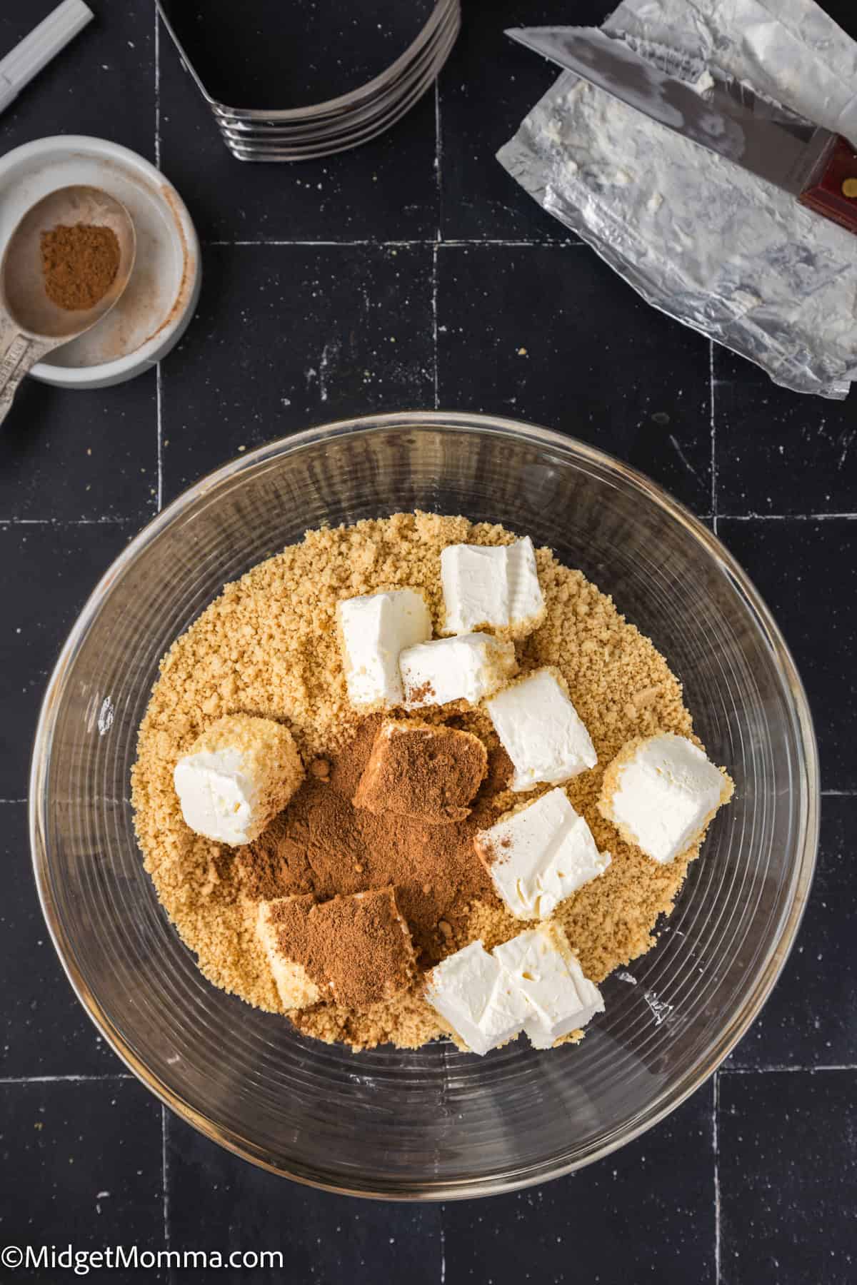 A glass bowl with crumbs, cubed butter, and brown sugar sits on a black counter next to a measuring spoon with spice and a wrapped block of butter.